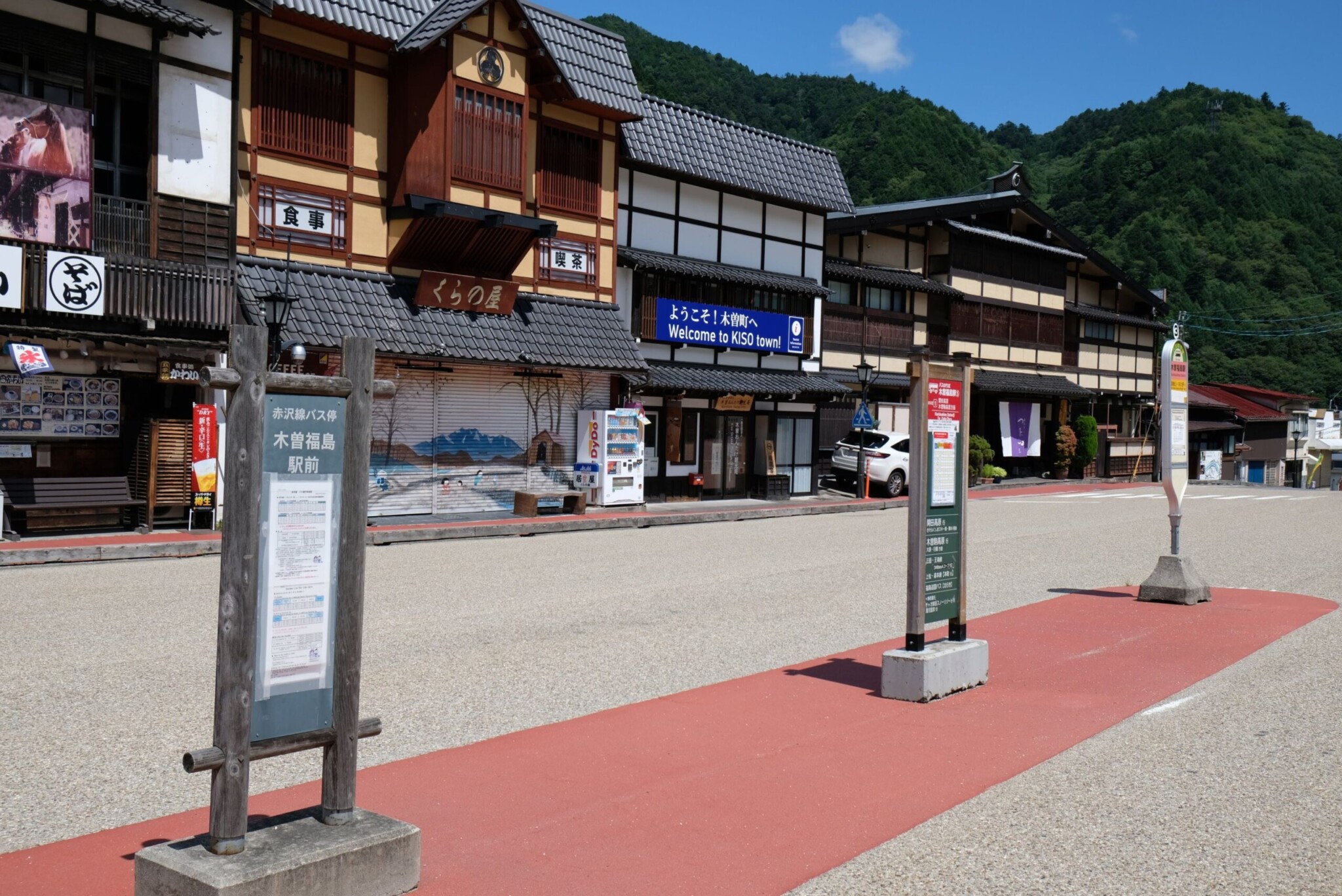 青空の下に並ぶ、木造風・和風建築の土産物店や食事処の店舗外観。右側の建物には白い壁に黒い梁が施され、中央の建物には黄色い日よけテントがある。手前は舗装された広場または道路になっており、赤く塗装されたレーンと白の横断歩道のような白線が見える。