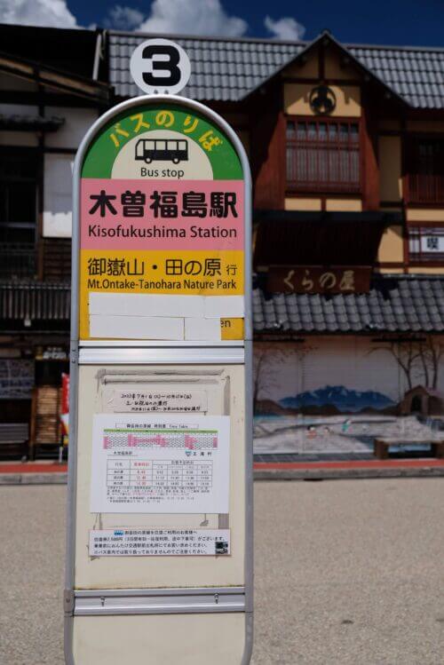 和風建築の駅前商店街が並ぶエリア。手前にはアスファルトの広場があり、赤く塗られたエリアに木製のバス停の看板や案内板が立っている。奥には「くらの屋」などの店舗が並び、山並みと青空が見える。