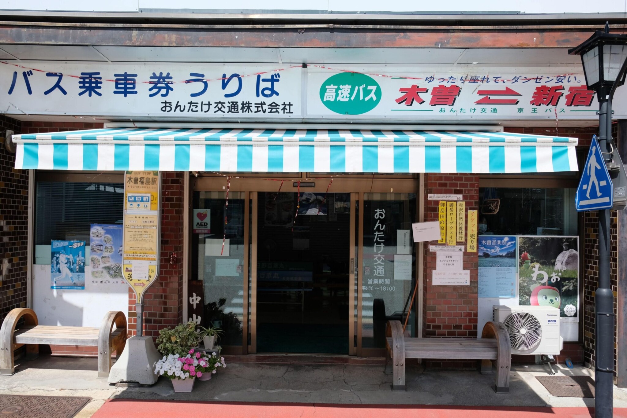 バス停留所のサイン(看板)のアップ。上部には「3」の数字があり、緑・ピンク・黄色のカラフルなデザインで路線案内が書かれている。下部には運行期間や時刻表、注意書きが書かれた紙が貼り付けられている。背景にはうっすらと「くらの屋」の建物がボケて写っている。