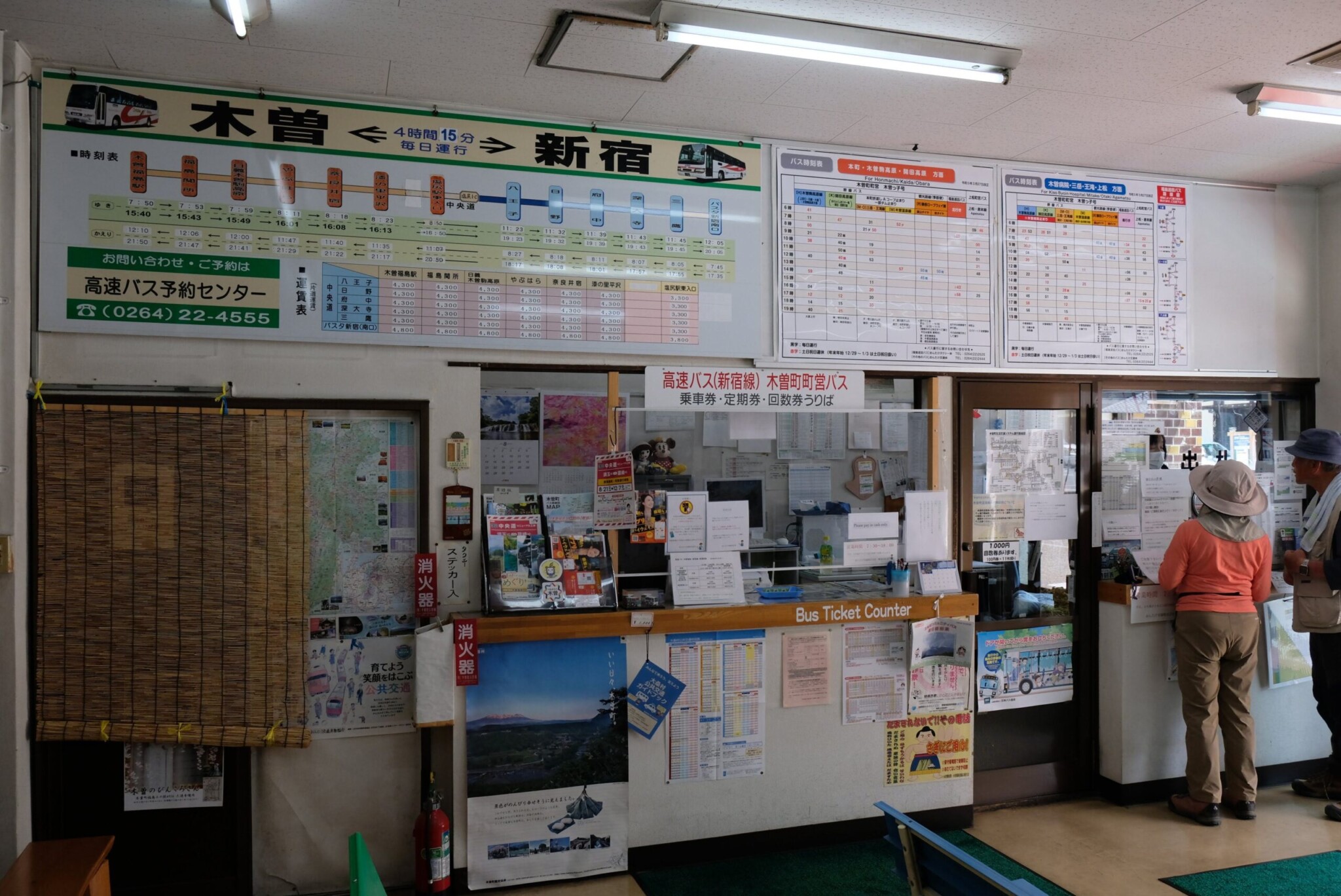 おんたけ交通株式会社の「バス乗車券うりば」の店舗正面。入り口はガラスの引き戸になっており、上部には青と白のストライプの日よけテントがある。入り口の左右には木製のベンチが置かれ、左側には植木鉢の花、右側にはエアコンの室外機がある。壁面には各種ポスターや案内が多数貼られている。