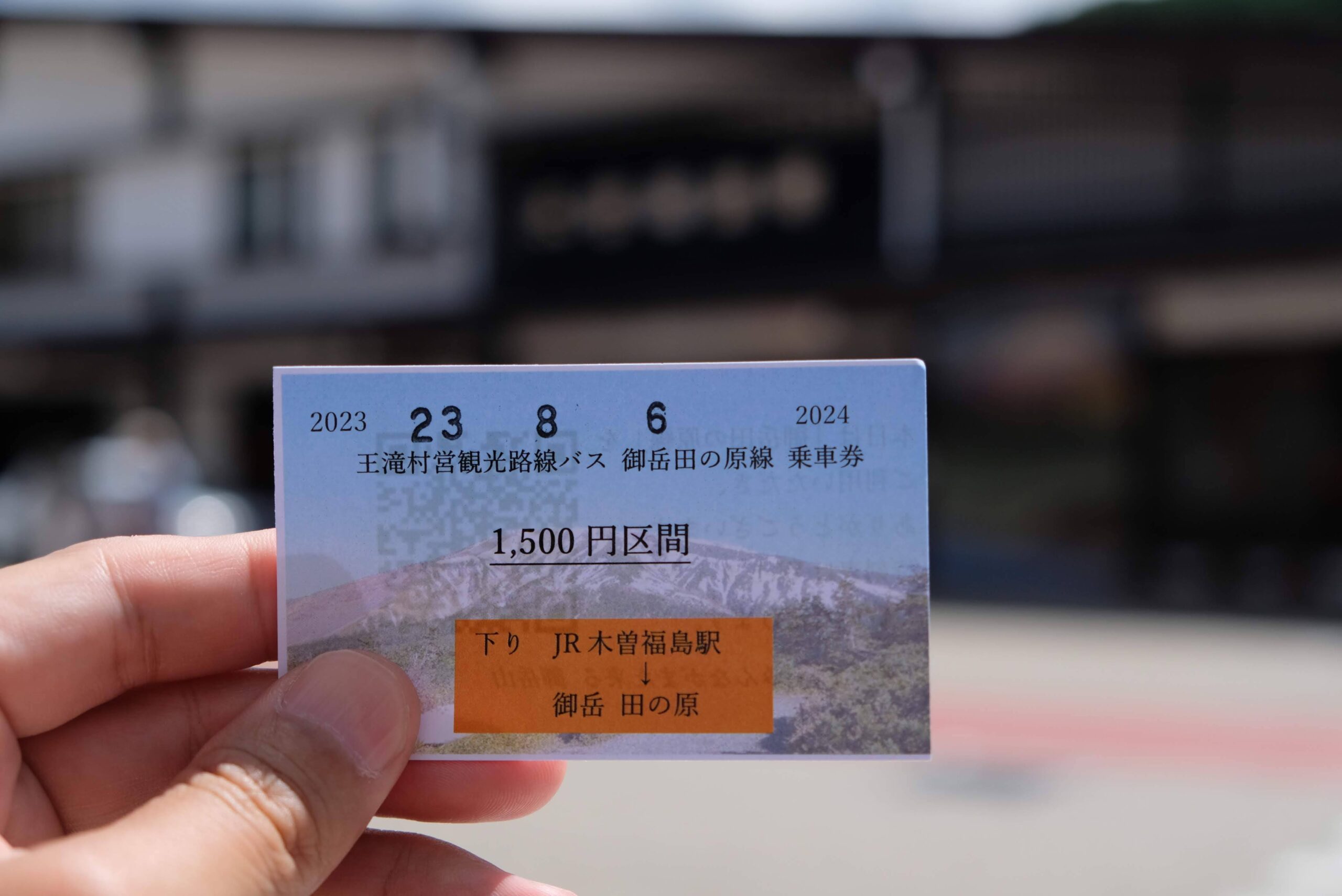 バス乗車券うりばの待合室・窓口の内部。木製のカウンター(Bus Ticket Counter)があり、その奥が事務スペースになっている。壁面には大きな路線図や高速バスの時刻表・料金表、各種ポスターが所狭しと貼られている。窓口の前にはリュックを背負い帽子をかぶった男性と女性(乗客)が立っており、案内を確認または購入している様子。