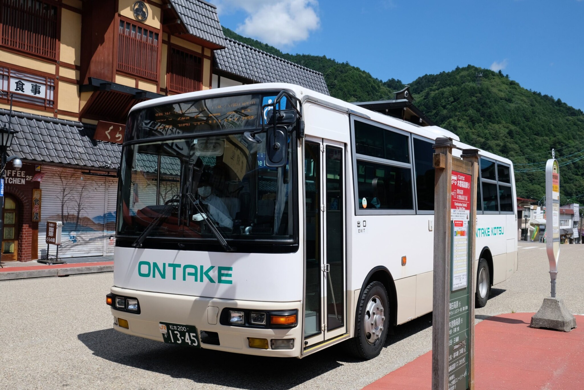 人物の手(親指と人差し指)で持たれた、バスの乗車券のクローズアップ。背景は木曽福島駅前の建物が大きくぼやけて写っている。切符には日付や路線名、金額が印字されており、背景にはうっすらと御嶽山のイラストが描かれている。