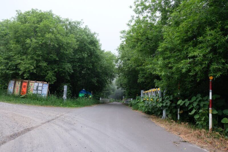 舗装路の終点付近、森の入り口に設置された複数の工事看板と斜里岳登山道への案内標識。