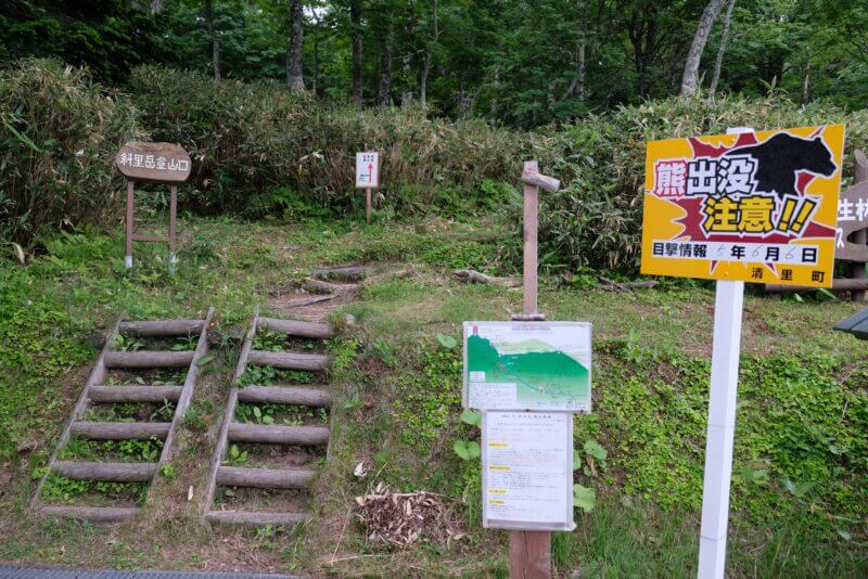 斜里岳登山口の入り口。熊出没注意の黄色い看板と登山コース案内、木製の標識が並ぶ。