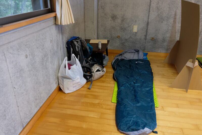 コンクリート壁の横に広げられた寝袋と登山用バックパック。壁には番号札が見える。