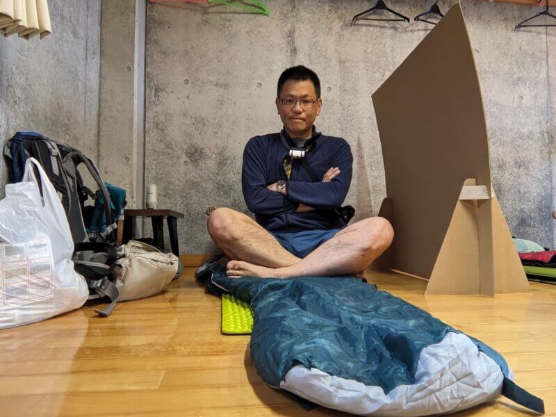 寝袋の上に座り、腕を組んでポーズをとる撮影者の自撮り写真。