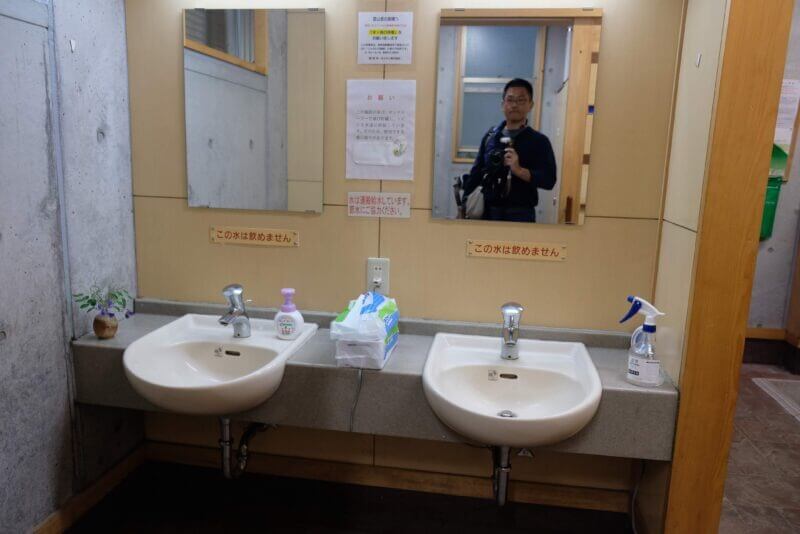 洗面台の鏡に写る撮影者。飲用不可と節水を促す多数の掲示物。