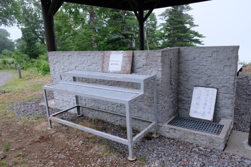 屋外のコンクリート製洗い場とザック置き場。泥落としの注意書きが貼られている。