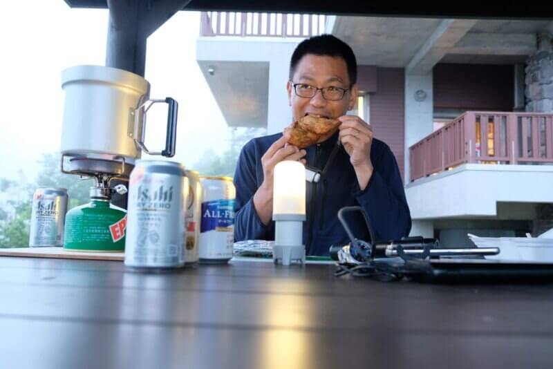 清岳荘のテラスで、ランタンの灯りの下、骨付き肉を食べる男性と並んだノンアルコールビール缶。