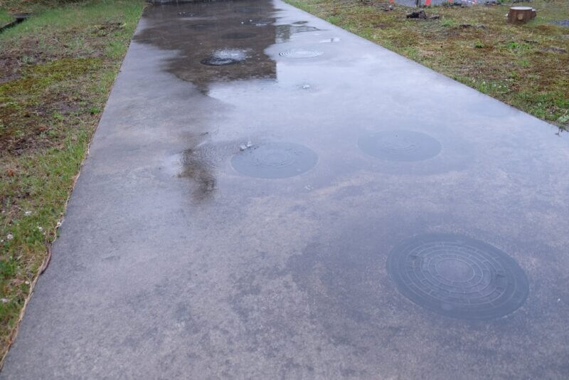 雨で濡れたコンクリートの通路。水溜まりに雨粒が落ちて波紋が広がっている。