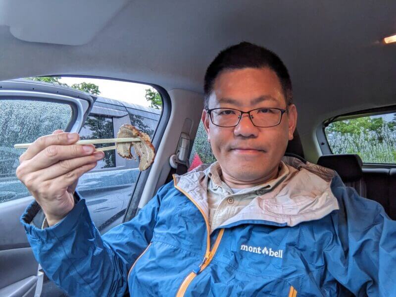 雨の車内でレインウェアを着たまま肉を食べる男性。