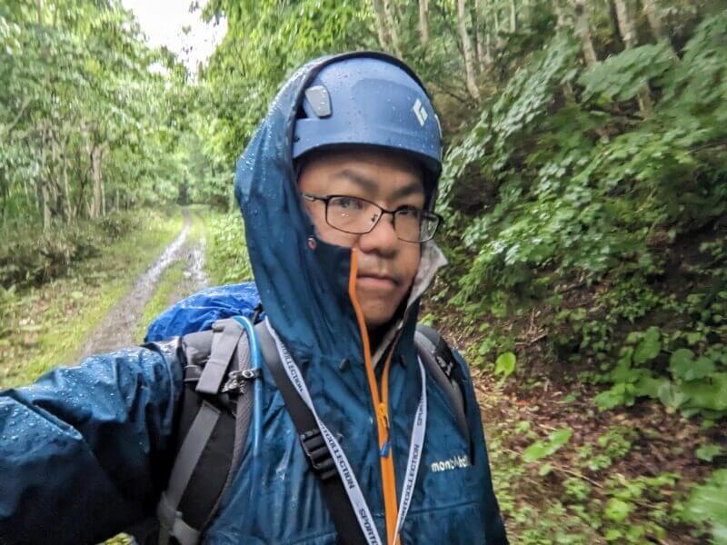 雨の中、青いヘルメットとレインウェアを着用して自撮りする執筆者の姿。眼鏡が濡れている。