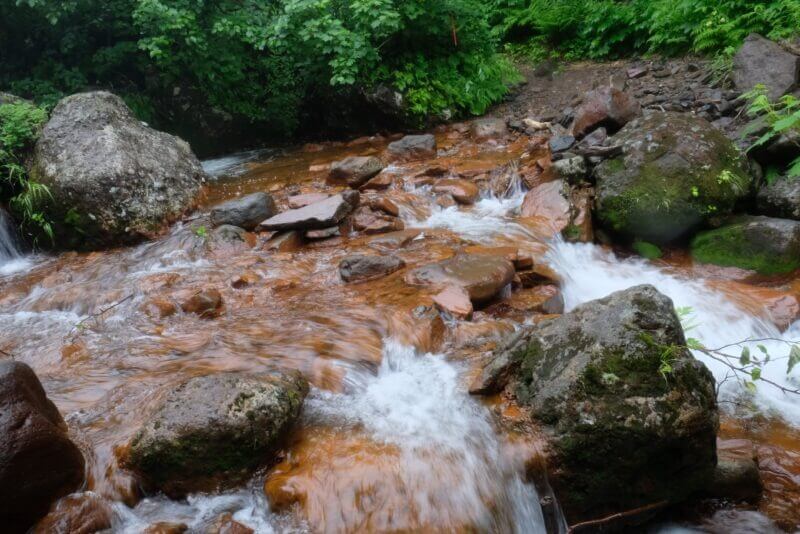 赤褐色の大きな岩が乱立する間を、激しく飛沫を上げて流れる沢の様子。
