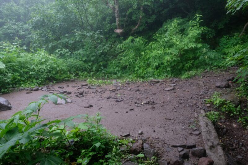 霧で視界が遮られた山中の平坦な場所。泥濘んだ地面に小石が散らばっている。