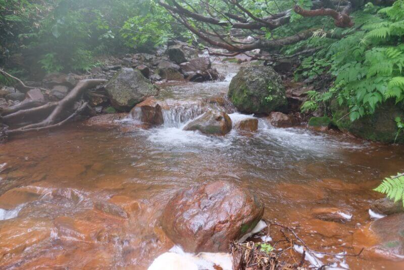 赤褐色の岩が重なる沢を、小さな滝となって流れ落ちる透明な水流。