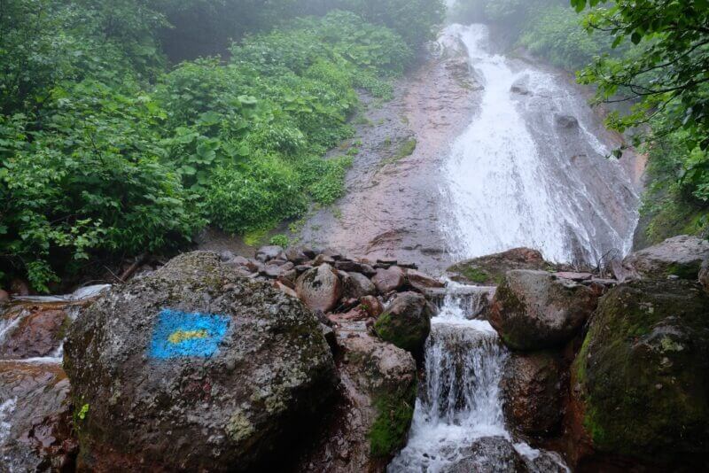 巨大な一枚岩を水が流れ落ちるナメ滝. 手前の岩には青地に黄色の矢印が描かれている。