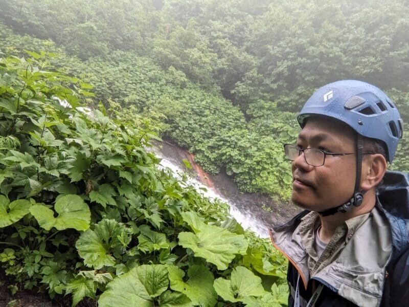 霧の深い登山道でブルーのヘルメットを被り自撮りをする筆者。周囲には大きなフキの葉が茂る。