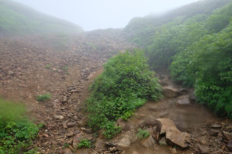 霧に包まれた茶褐色の広大なガレ場と登山道の斜面