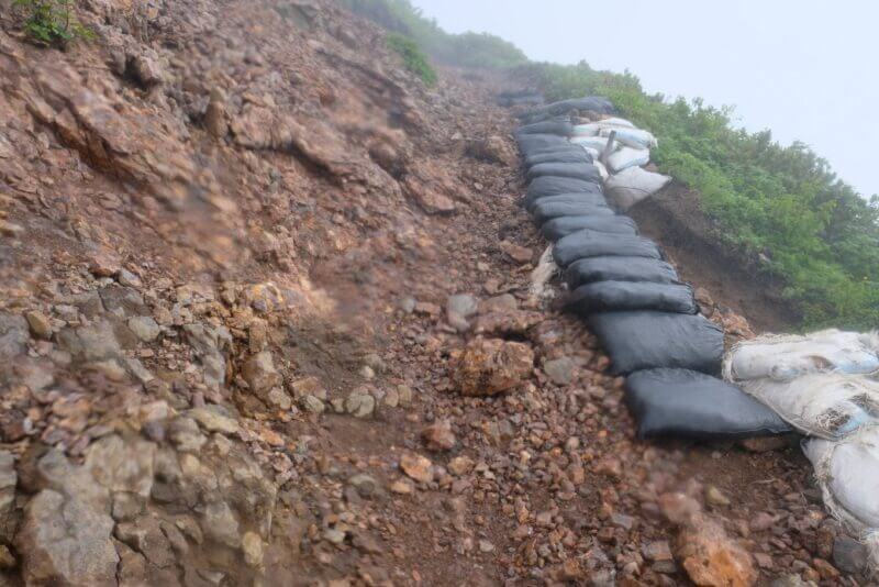 斜面の崩落を防ぐために黒い土嚢が並べられた急な登山道