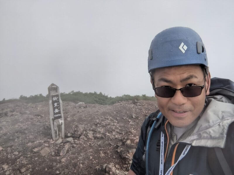 斜里岳山頂標識の横でヘルメットとサングラス姿で自ドリする男性