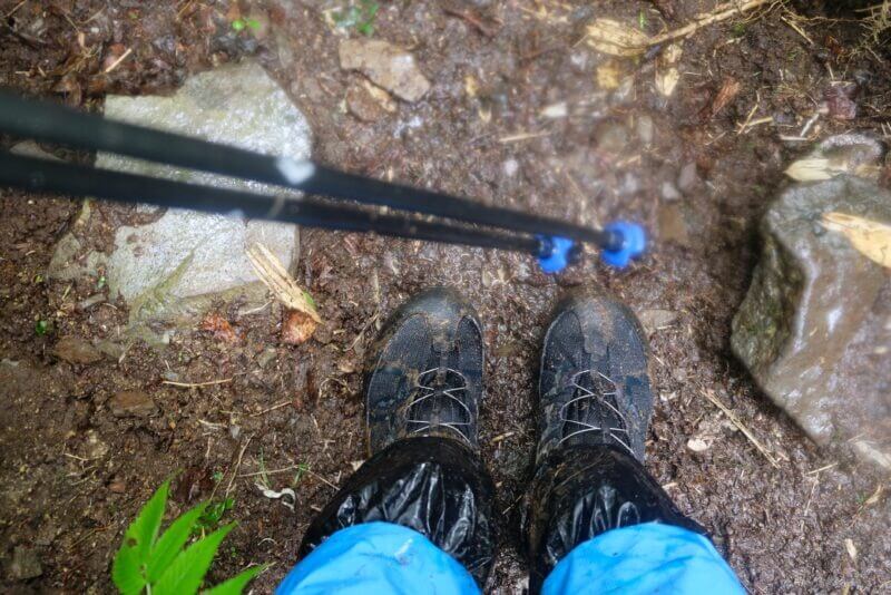 泥だらけの登山道を歩く登山者の足元。黒い登山靴と青いレインウェア,ストックが見える。