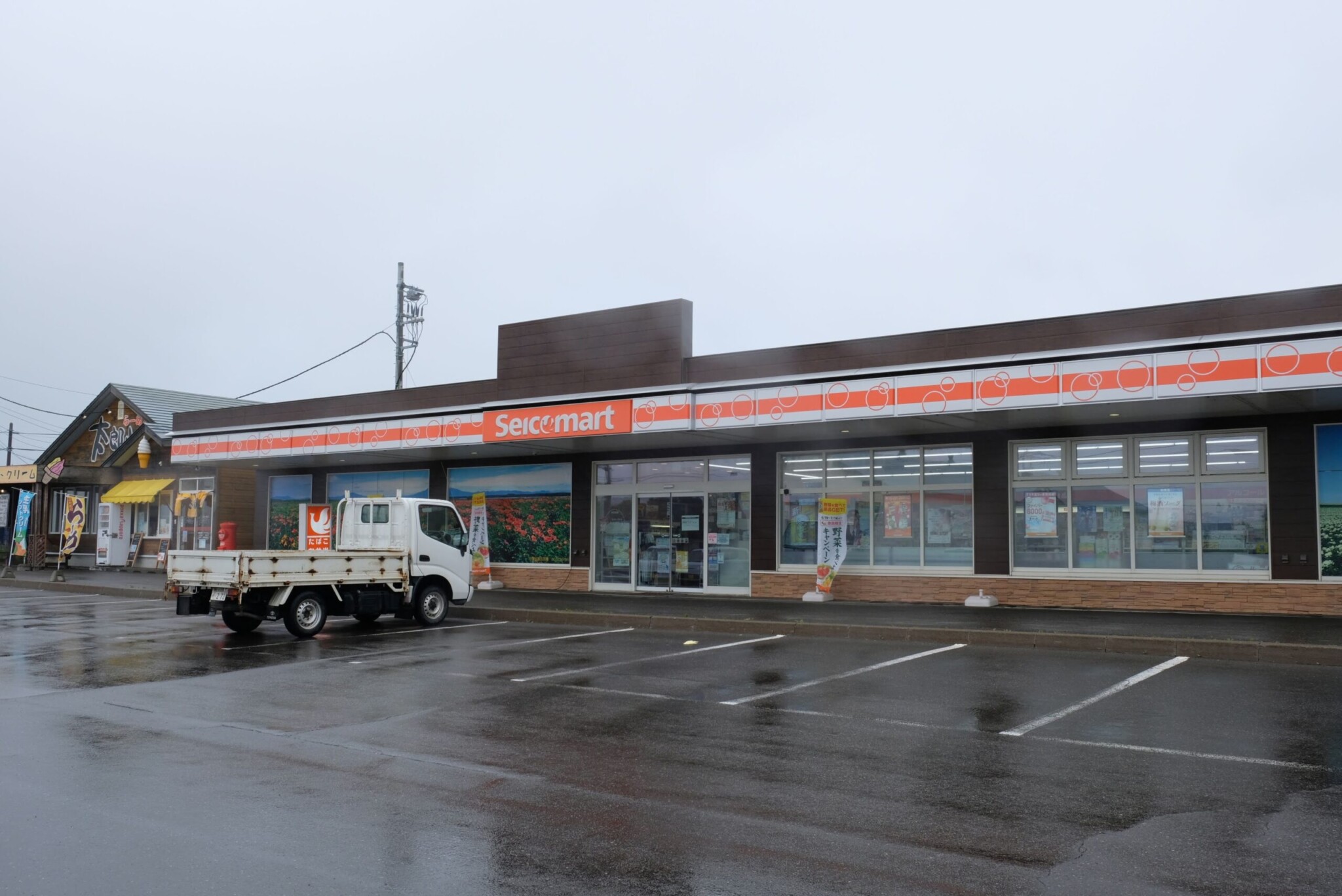 雨の中のセイコーマート小清水道の駅店。駐車場が雨に濡れて反射している。