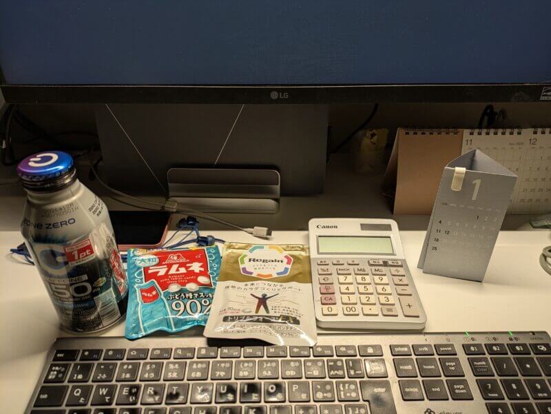 PCモニターの前に並べられたZONe ENERGY、森永ラムネ、リゲインのサプリメント、キヤノンの電卓、卓上カレンダーが置かれたデスク周りの様子